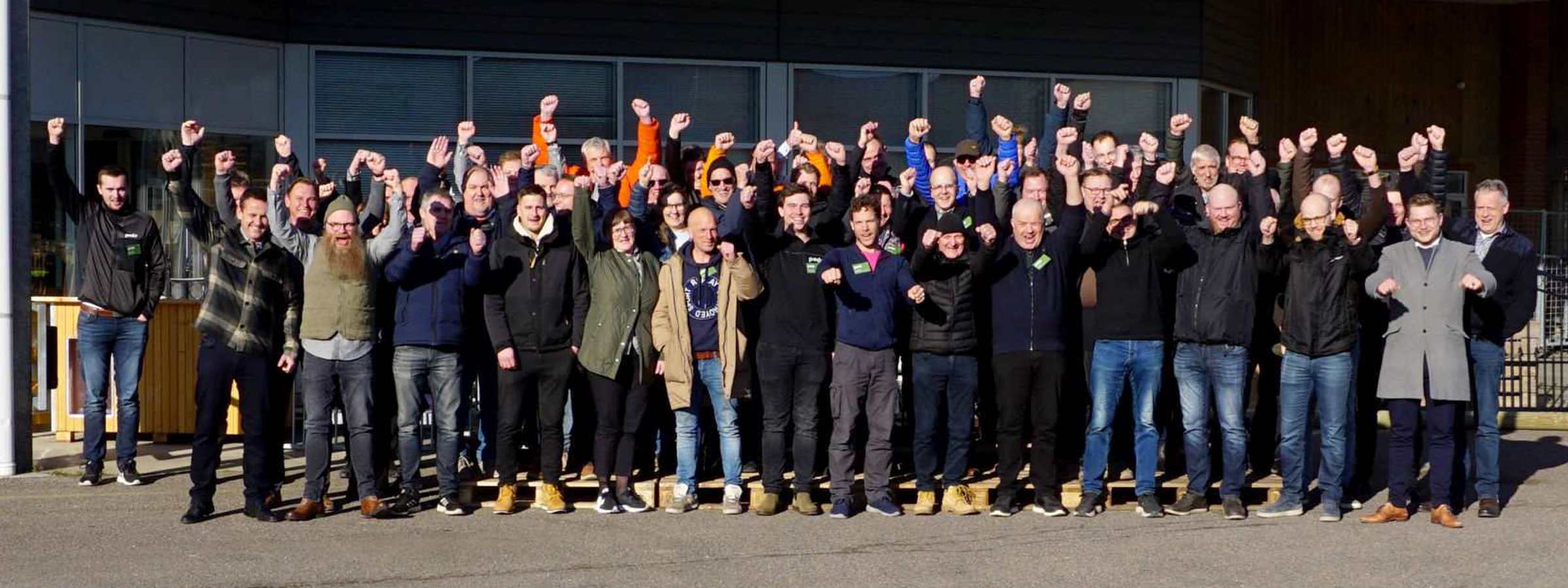 Gruppefoto af Poda-medarbejdere foran virksomhedens hovedbygning med Poda-logo og hævede arme i fællesskab.