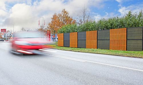 Noistop Essential støjhegn med sort ramme og metalpaneler, hvor nogle sektioner har tilvalgte træpaneler monteret udenpå. Hegnet er omgivet af grøn beplantning, og en rød bil passerer hurtigt forbi på vejen i forgrunden.