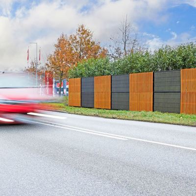Noistop Essential støjhegn med sort ramme og metalpaneler, hvor nogle sektioner har tilvalgte træpaneler monteret udenpå. Hegnet er omgivet af grøn beplantning, og en rød bil passerer hurtigt forbi på vejen i forgrunden.