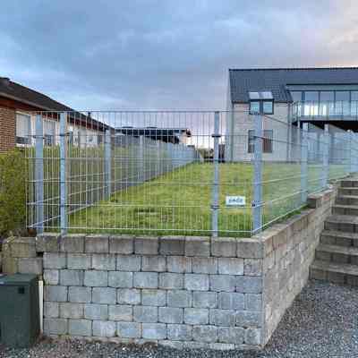 Nærbillede af et panelhegn monteret på en forhøjet græsplæne med støttemur af betonblokke. I baggrunden ses et moderne hus med store glaspartier.