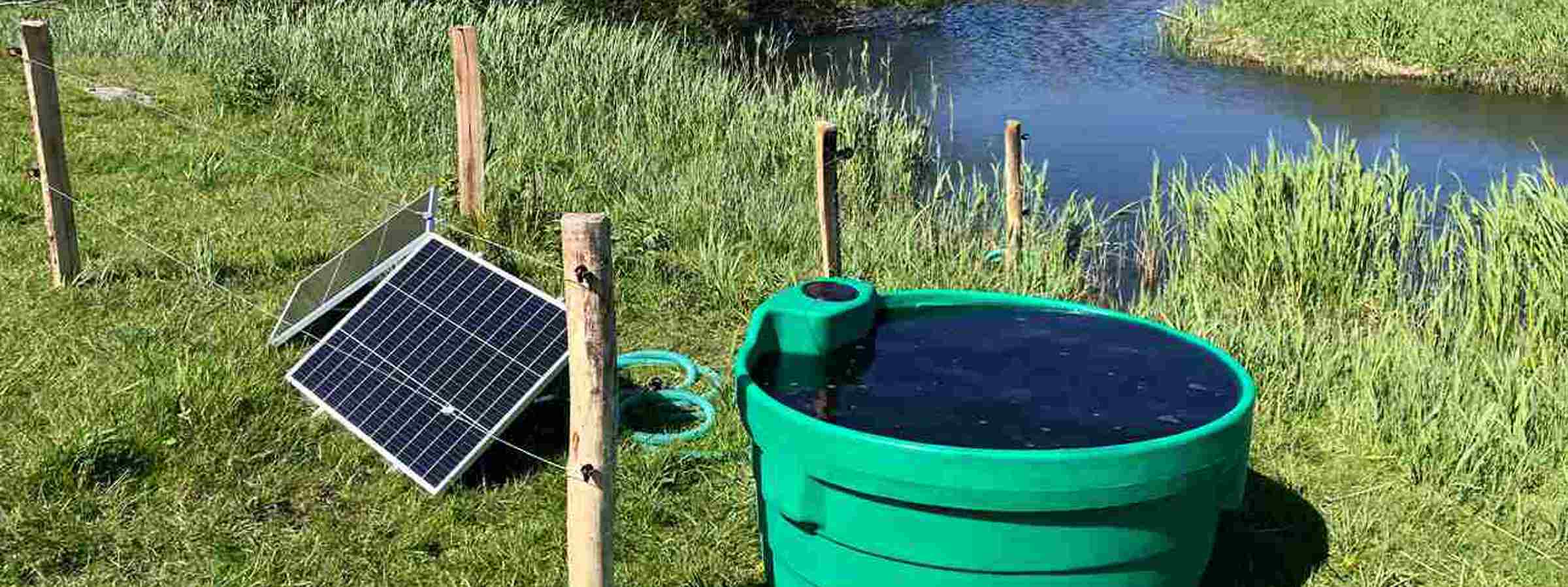 Soldrevet markpumpe med solpanel og vandkar ved en å på en græsmark