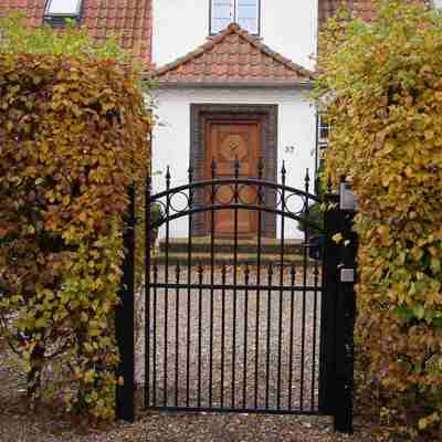 En ganglåge i sort smedejern er monteret mellem to buske som udgør indgangen til et hus