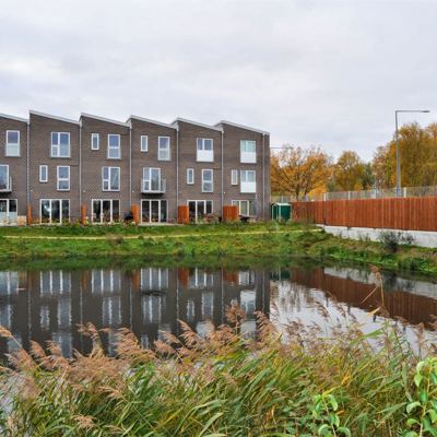 Et træbeklædt Noistop® Steel støjhegn fra Poda Hegn opsat foran et grønt område med en lille sø ved et rækkehusområde til afskærmning af trafikstøj fra en større offentlig vej