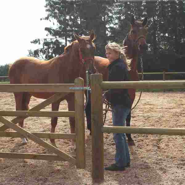 En dame står, sammen med sine to heste, bag et lægtehegn og en Country Farmer låge