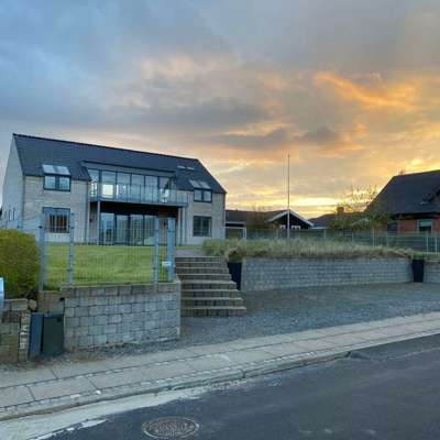 Moderne to-etagers hus med store glaspartier og balkon, omgivet af et panelhegn. Huset ligger på en hævet grund med trapper op til haven og en flot solnedgang i baggrunden.