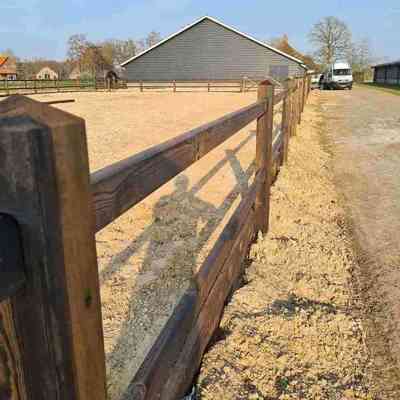 En ridebane afgrænses af et elegant hestehegn. I baggrunden ses flere staldbygninger.