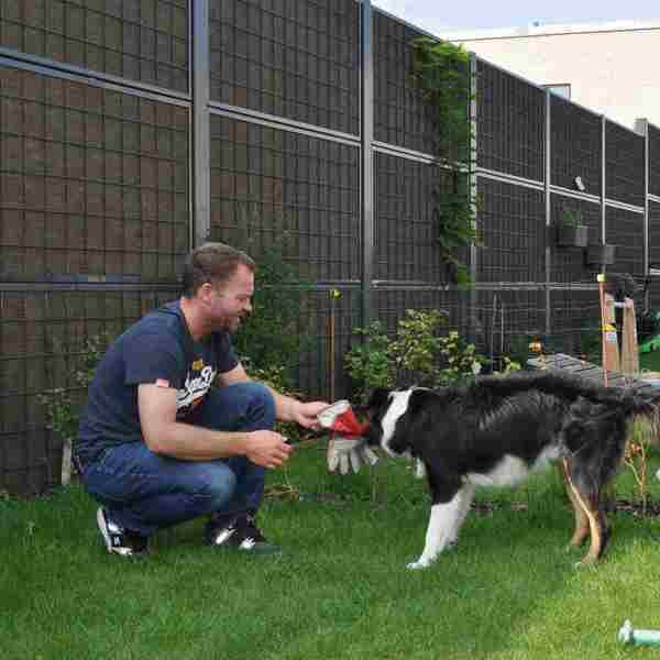En mand og hans hund sidder foran et Noistop® Steel støjhegn fra Poda Hegn der er opsat i en have til afskærmning af støj fra naboerne