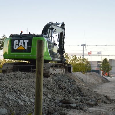 På en byggeplads er en grøn CAT-maskine parkeret. For at øge sikkerheden på pladsen, er der monteret et byggepladshegn