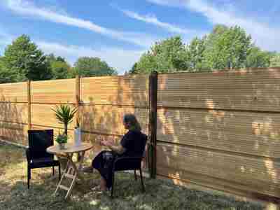 En kvinde sidder ved et bord med en plante og en kande foran et DeciFence træstøjhegn på en solrig dag.