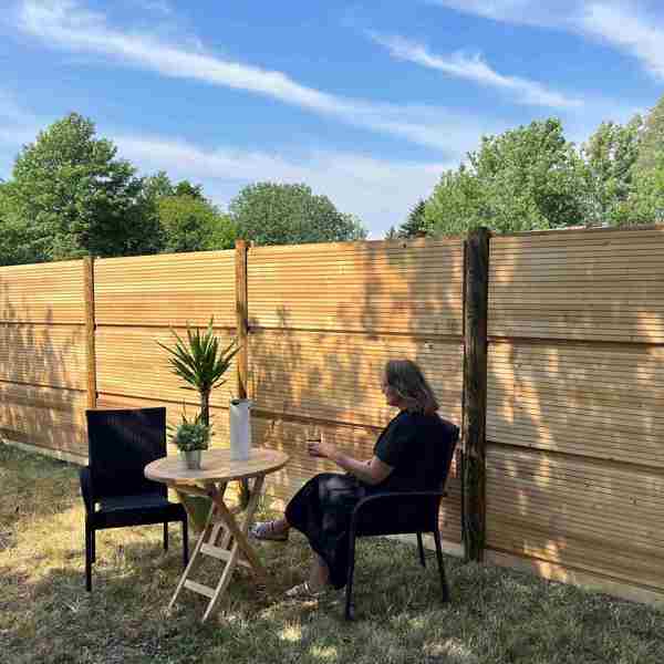 En kvinde sidder ved et bord med en plante og en kande foran et DeciFence træstøjhegn på en solrig dag.