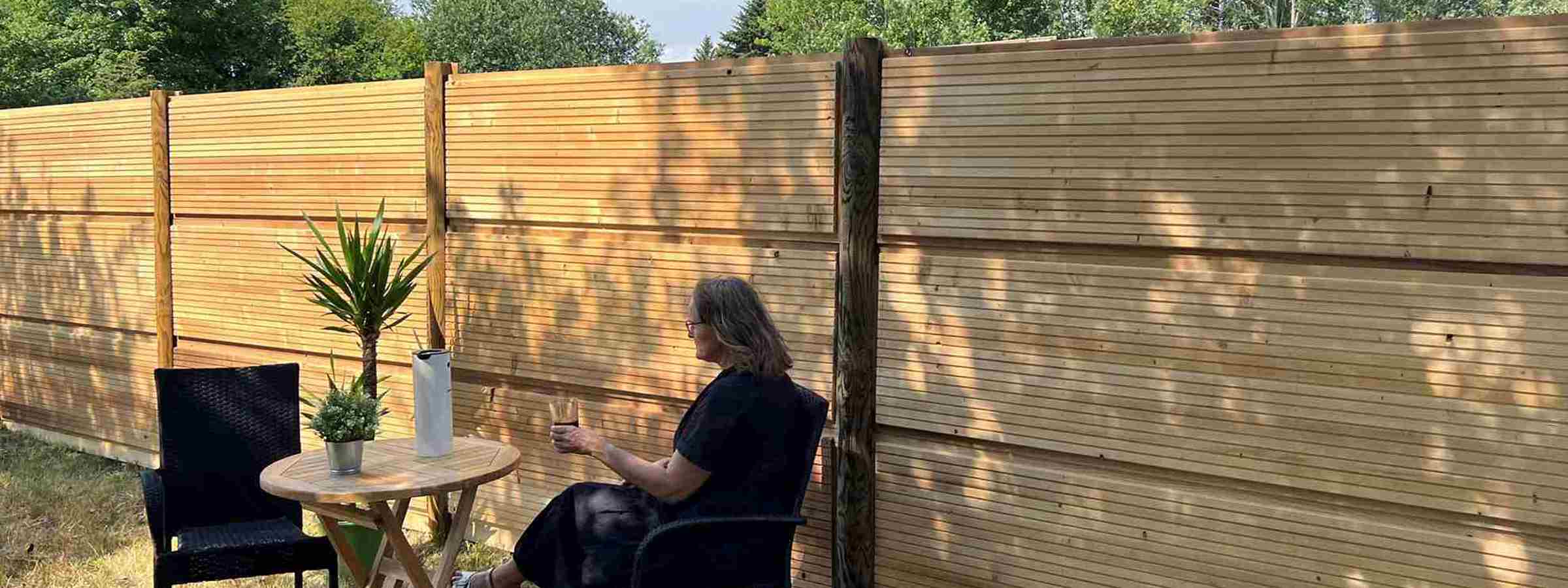 En kvinde sidder ved et bord med en plante og en kande foran et DeciFence træstøjhegn på en solrig dag.