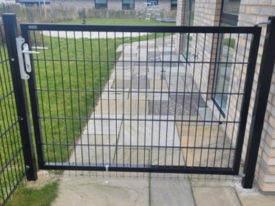 Sort metal låge med panelhegn, monteret mellem en murstensbygning og en sort metalstolpe. Lågen har et metalhåndtag og en lås, og leder til en stenbelagt gangsti med græs på begge sider.