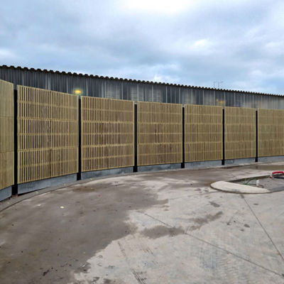 Buet Noistop Essential støjhegn med sort ramme og lyse trælameller, monteret på betonfod i et industriområde.