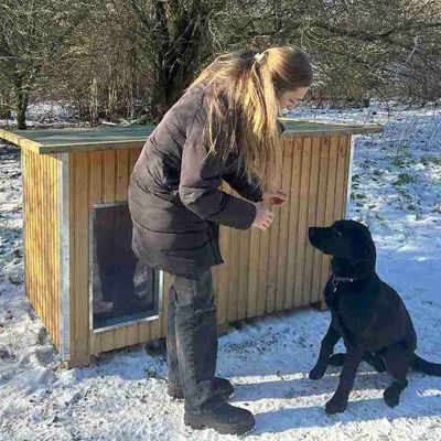 Kvinde giver en sort hund en godbid foran et Luksus hundehus i et vinterlandskab med sne på jorden.