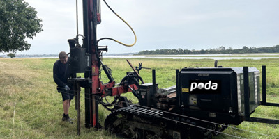 En montør banker pæle ned med en pælehammer monteret på bæltekøretøj i et åbent landskab nær vandet.