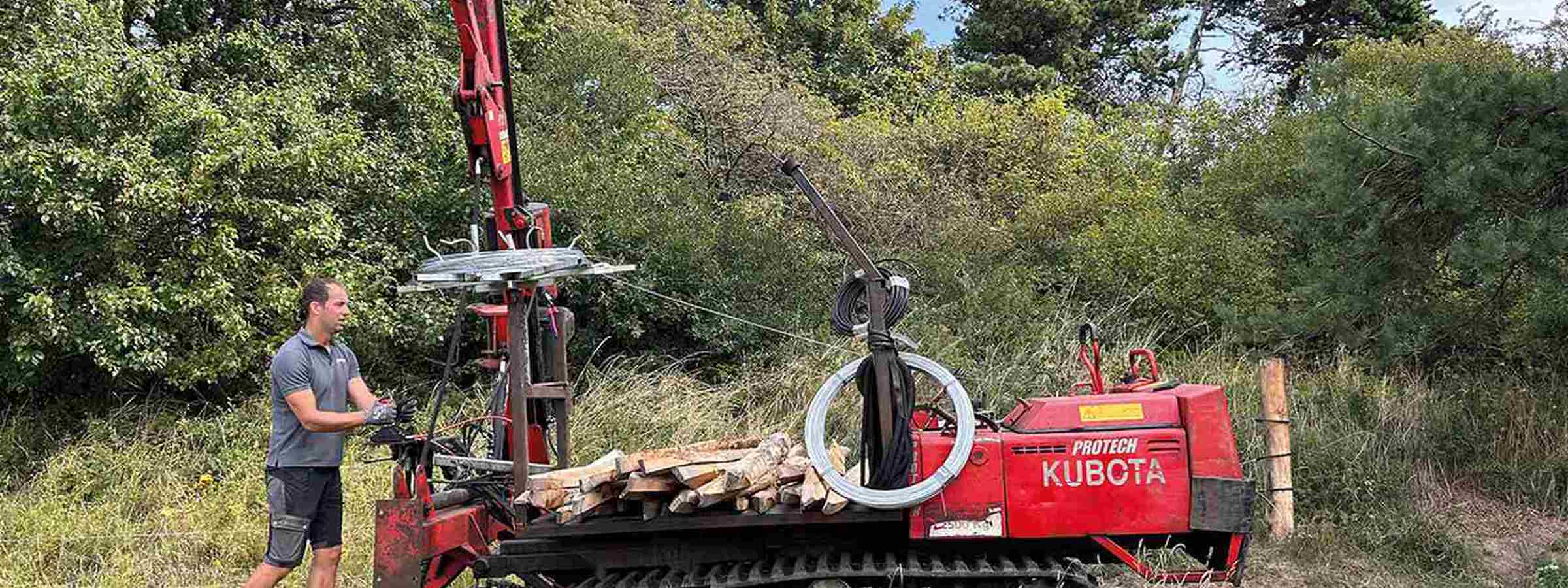 En montør er i gang med at rulle tråd ud til et elektrisk hegn med en specialbygget maskine i et grønt og skovrigt område.