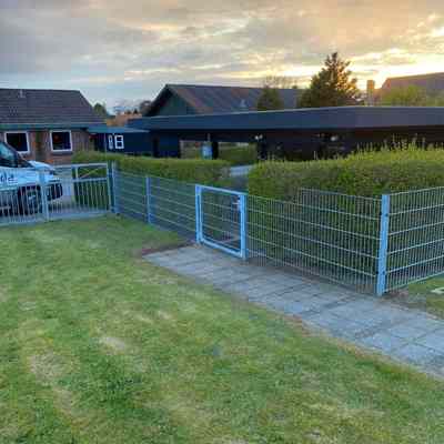 Panelhegn med en låge monteret langs en sti, der adskiller en græsplæne fra en hæk. I baggrunden ses en carport, flere huse og en solnedgang.