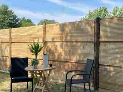 DeciFence støjhegn med lodrette træstolper og vandrette paneler. Foran hegnet står et cafésæt med to sorte stole, et lille rundt træbord, en plante og en termokande.