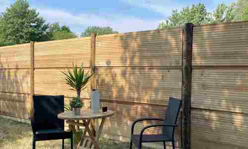 DeciFence støjhegn med lodrette træstolper og vandrette paneler. Foran hegnet står et cafésæt med to sorte stole, et lille rundt træbord, en plante og en termokande.