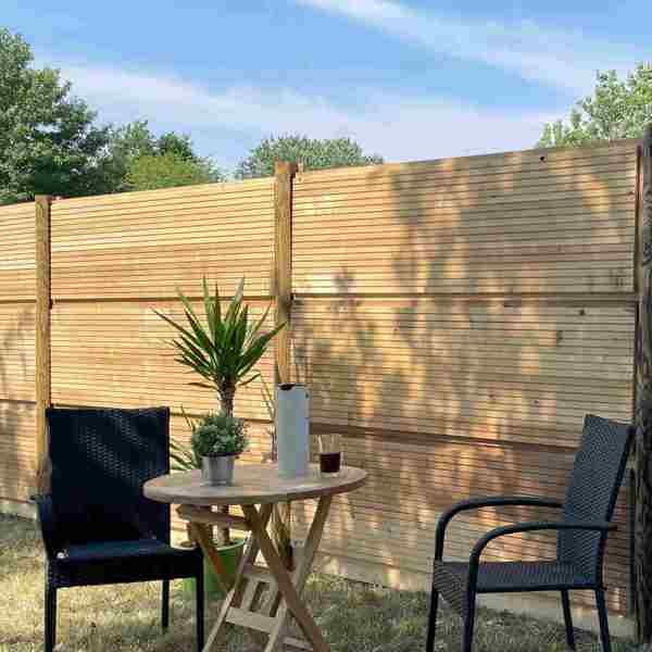 DeciFence støjhegn med lodrette træstolper og vandrette paneler. Foran hegnet står et cafésæt med to sorte stole, et lille rundt træbord, en plante og en termokande.