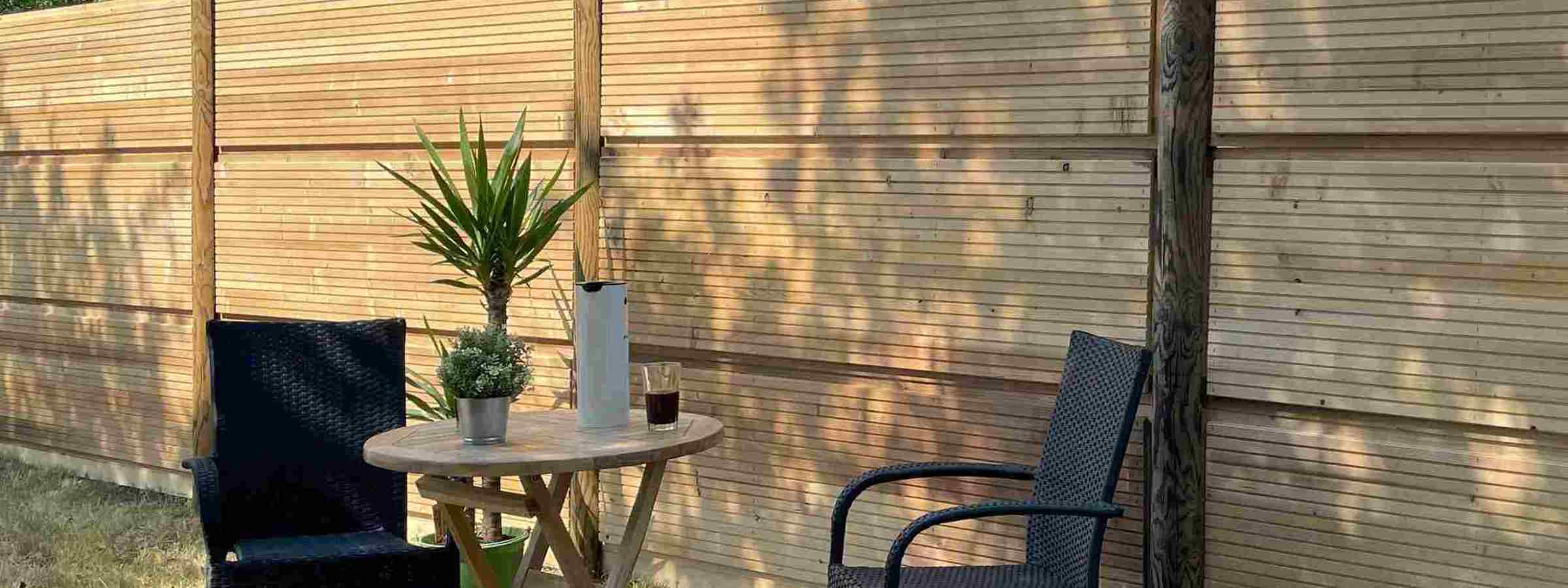 DeciFence støjhegn med lodrette træstolper og vandrette paneler. Foran hegnet står et cafésæt med to sorte stole, et lille rundt træbord, en plante og en termokande.